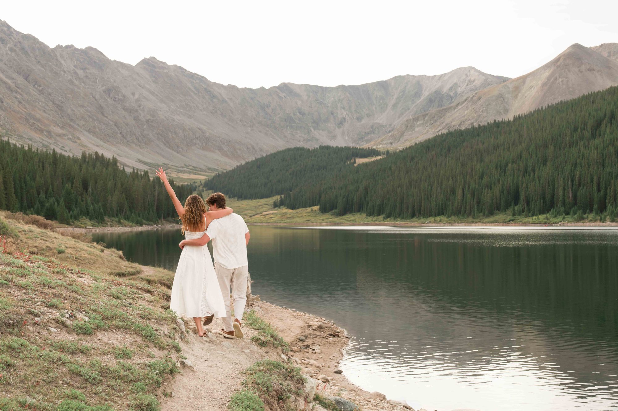 Anna raising her hand to show her ring as she and Brock walk along a mountain lake